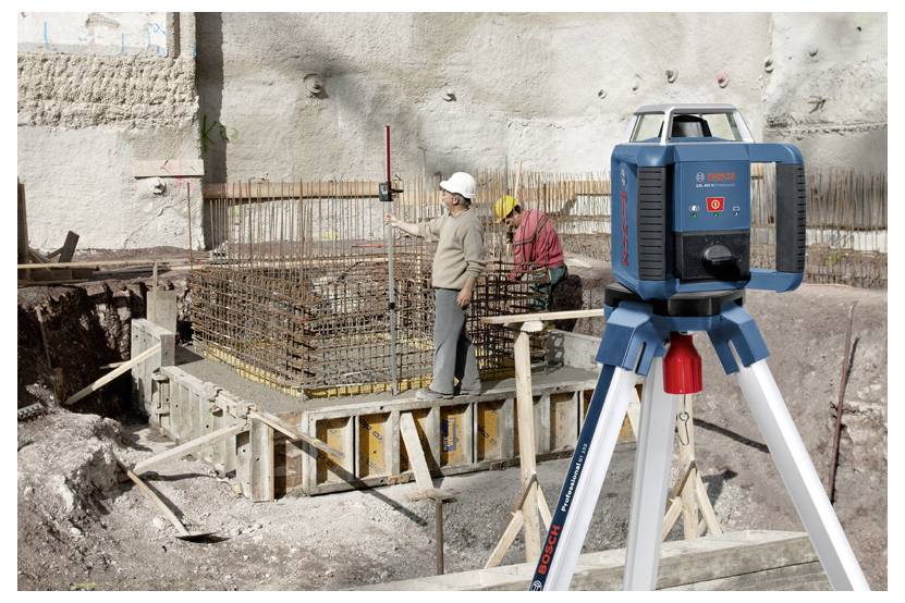 Baustelle mit einem Lasernivelliergerät auf einem Stativ im Vordergrund. Zwei Arbeiter mit Schutzhelmen arbeiten an einer verstärkten Stahlkonstruktion.