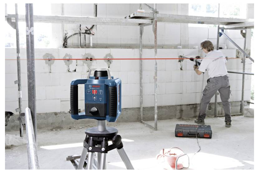 Ein Arbeiter verwendet ein Elektrowerkzeug, um in eine Ziegelwand auf einer Baustelle zu bohren. Ein Linienlaser befindet sich im Vordergrund und projiziert eine rote Linie.