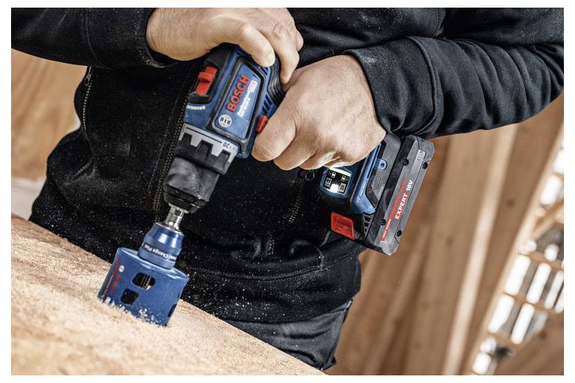 Eine Person, die einen Bosch-Akku-Bohrer mit einem Holzlochbohrer-Aufsatz verwendet, um in ein Stück Holz zu bohren und dabei Sägespäne zu erzeugen.