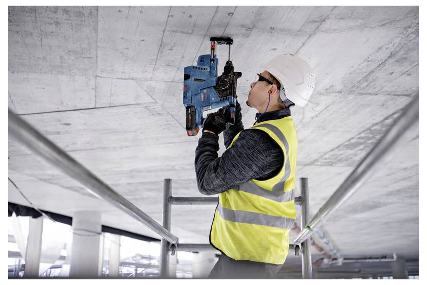 Ein Bauarbeiter mit Schutzhelm und Warnweste benutzt ein Elektrowerkzeug, um eine Betondecke beim Arbeiten auf einem Gerüst zu bohren.