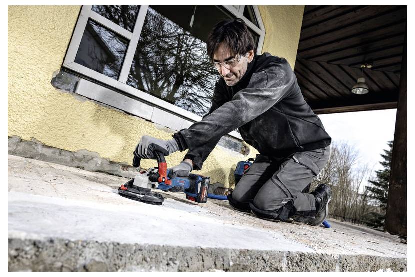 Eine Person in Schutzausrüstung kniet auf einer Betonoberfläche und verwendet eine elektrische Trennschleifmaschine zur Oberflächenvorbereitung neben einer Gebäudewand.