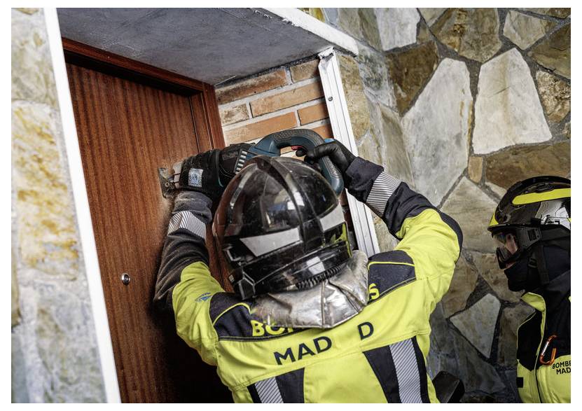 Zwei Feuerwehrleute in Schutzausrüstung schneiden eine Holztür in einem Steinmauernraum durch, mit Fokus auf dem aktiv arbeitenden Feuerwehrmann.