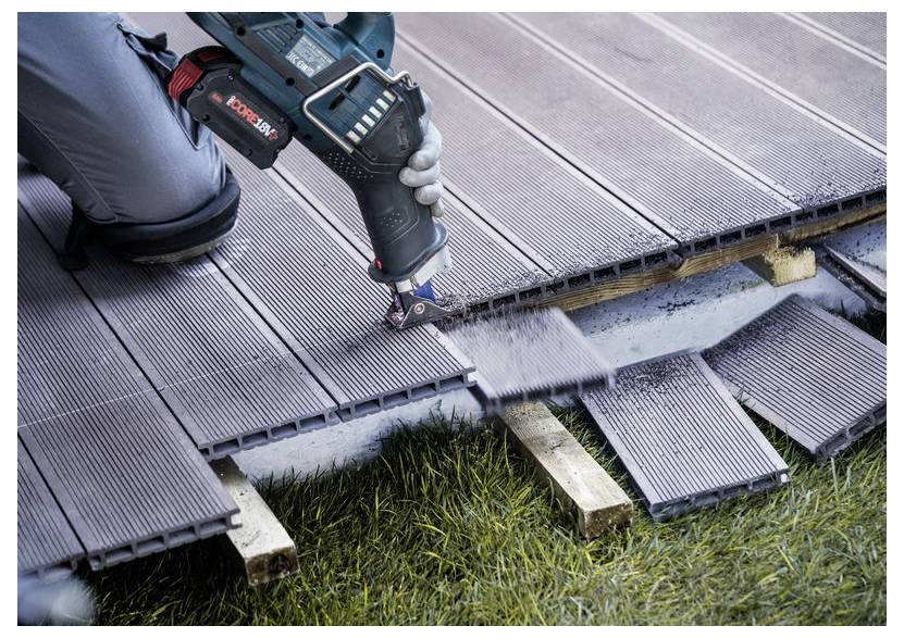 Eine Person, die ein Elektrowerkzeug verwendet, um graue Verbundwerkstoff-Terrassendielen über eine unebene Grasfläche zu schneiden.