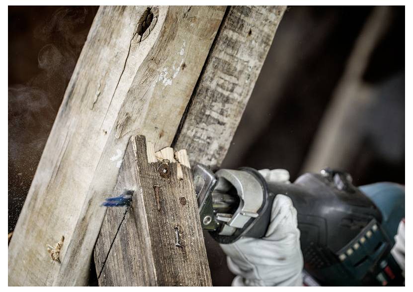 Eine Person mit Handschuhen verwendet eine Kreissäge, um ein altes, verwittertes Holzstück zu zersägen und dabei Sägespäne und Rauch zu erzeugen.