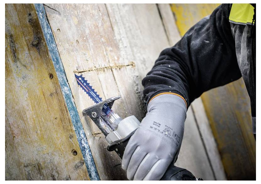 Eine Person in Schutzhandschuhen verwendet eine Säbelsäge mit blauer Klinge, um ein Holzbrett zu durchtrennen, wobei Sägespäne sichtbar sind.