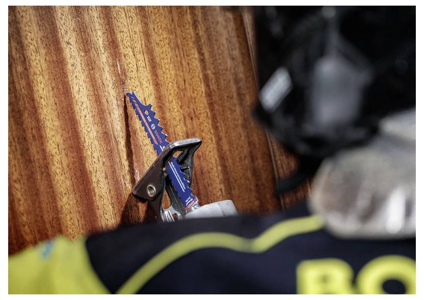 Eine blaue Sägeblatt ist an einer Holzoberfläche geklemmt, wobei eine Person mit Schutzausrüstung teilweise im Vordergrund sichtbar ist.