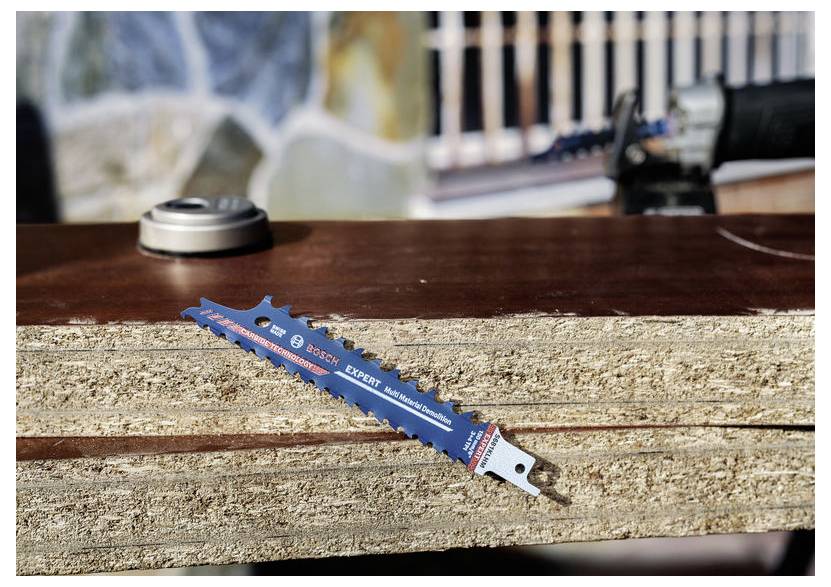 Eine Stichsägeblatt mit der Bezeichnung "Bosch Expert" liegt auf geschichteten Holzbrettern im Freien, während unscharfe Baumaterialien im Hintergrund zu sehen sind.