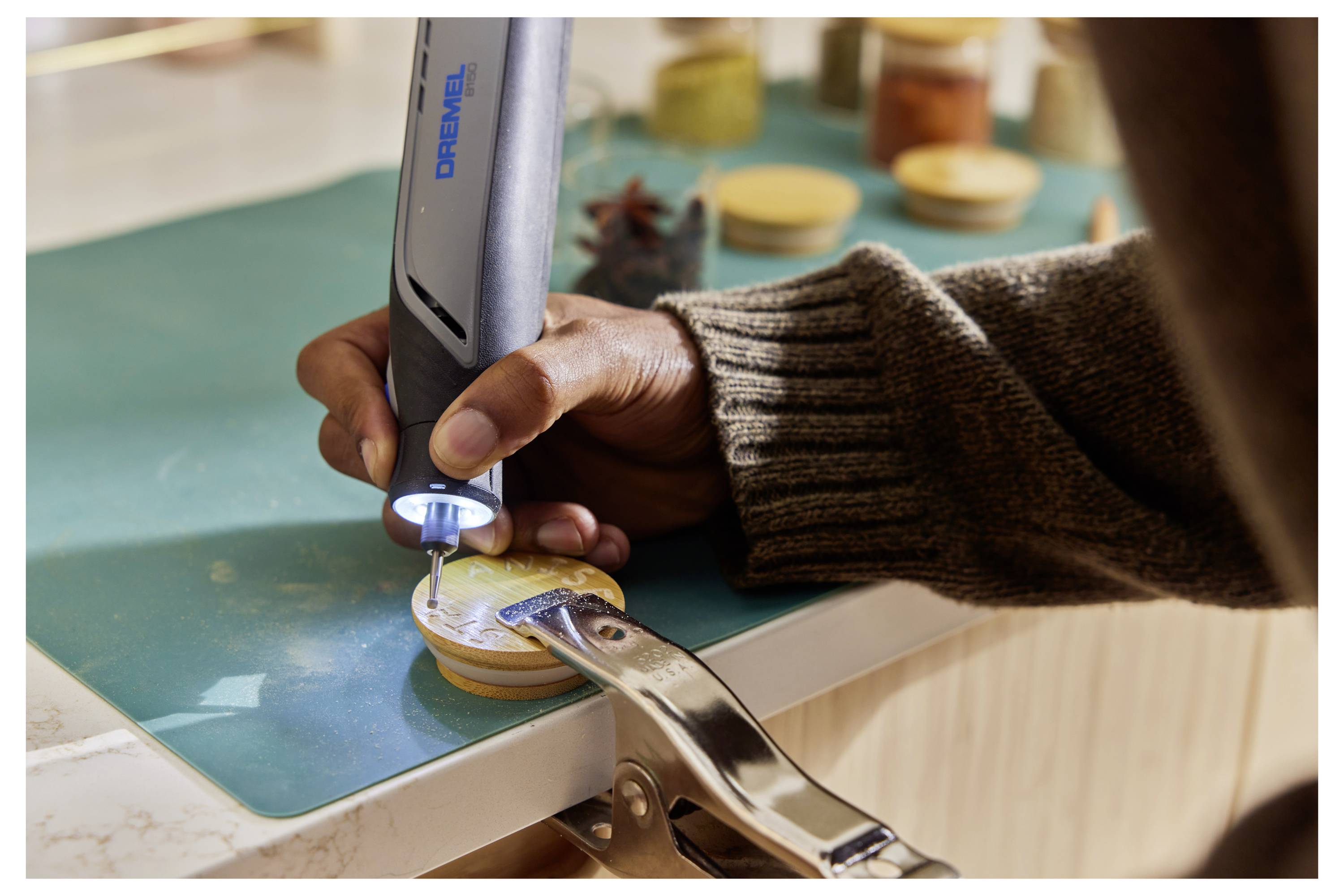 Eine Person verwendet ein tragbares Dremel-Werkzeug, um komplizierte Muster in ein kleines Holzobjekt zu schnitzen, das an einer Werkbank befestigt ist.