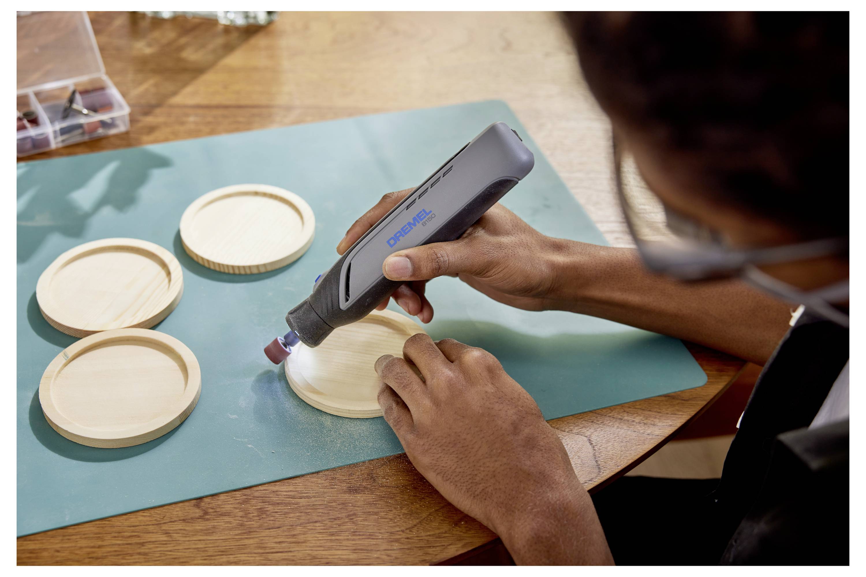 Eine Person benutzt ein tragbares Elektrowerkzeug, um runde Holzuntersetzer auf einem Tisch zu schleifen und sich dabei auf das Glätten der Oberfläche zu konzentrieren.