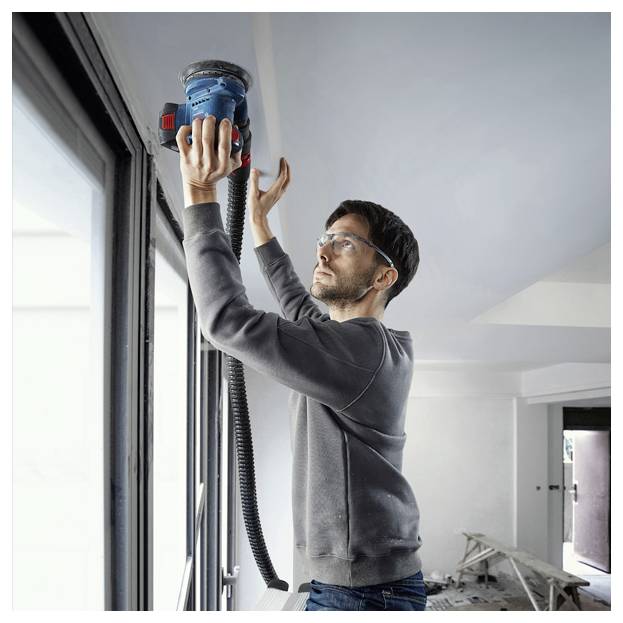 Eine Person mit Sicherheitsbrille benutzt einen blauen Exzenterschleifer an einer Decke in einem Raum, der renoviert wird, wobei Werkzeuge und Trümmer im Hintergrund sichtbar sind.
