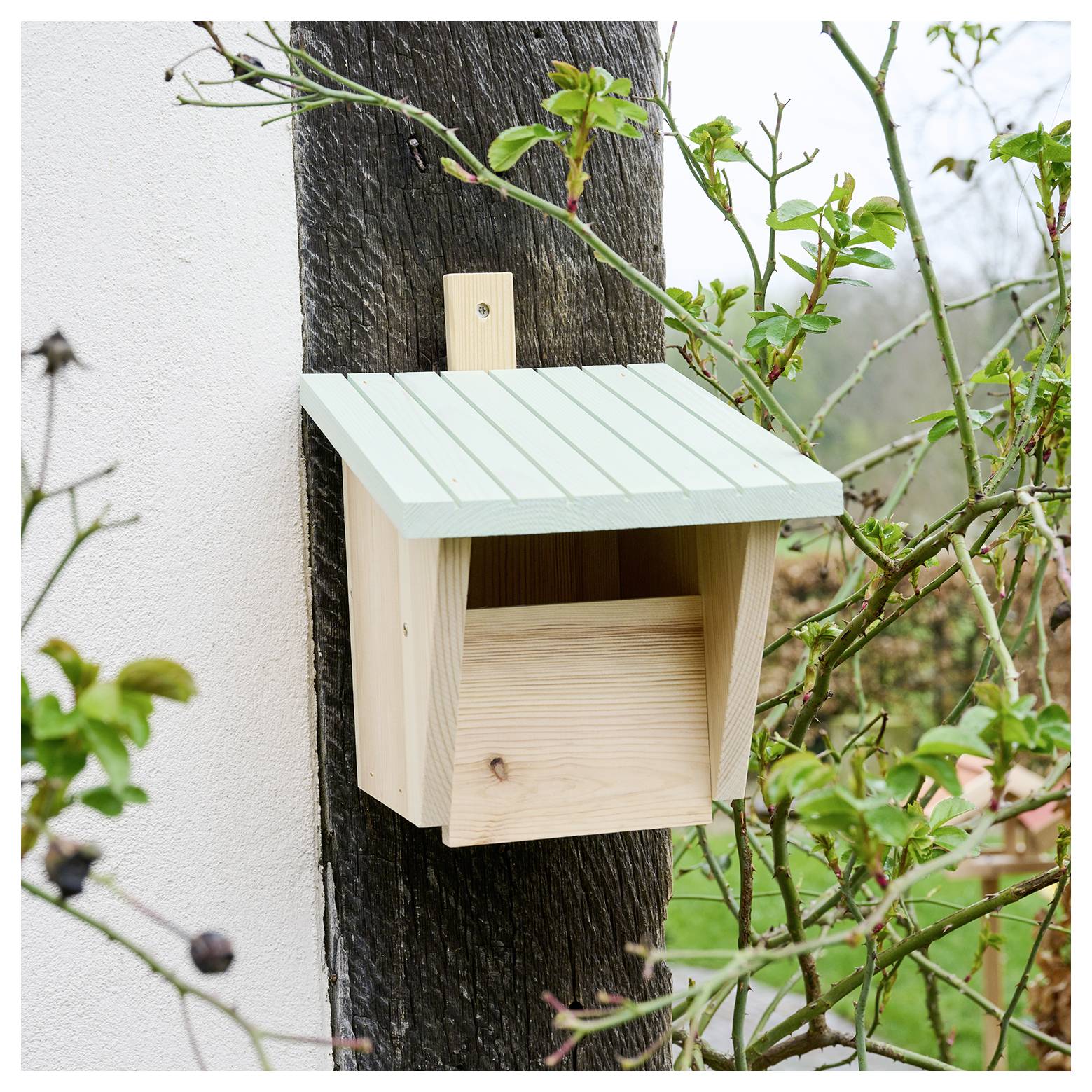 Ein kleines Holzvogelhäuschen mit einem grünen Dach ist auf einem Holzpfosten montiert und von grünem Laub umgeben.