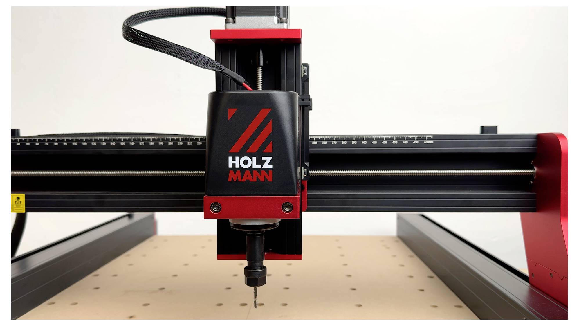 Eine CNC-Maschine mit einem 'Holzmann' Markenlabel, mit einem Bohrer, der über einer gelochten Arbeitsoberfläche positioniert ist, was auf präzise Bearbeitung hindeutet.