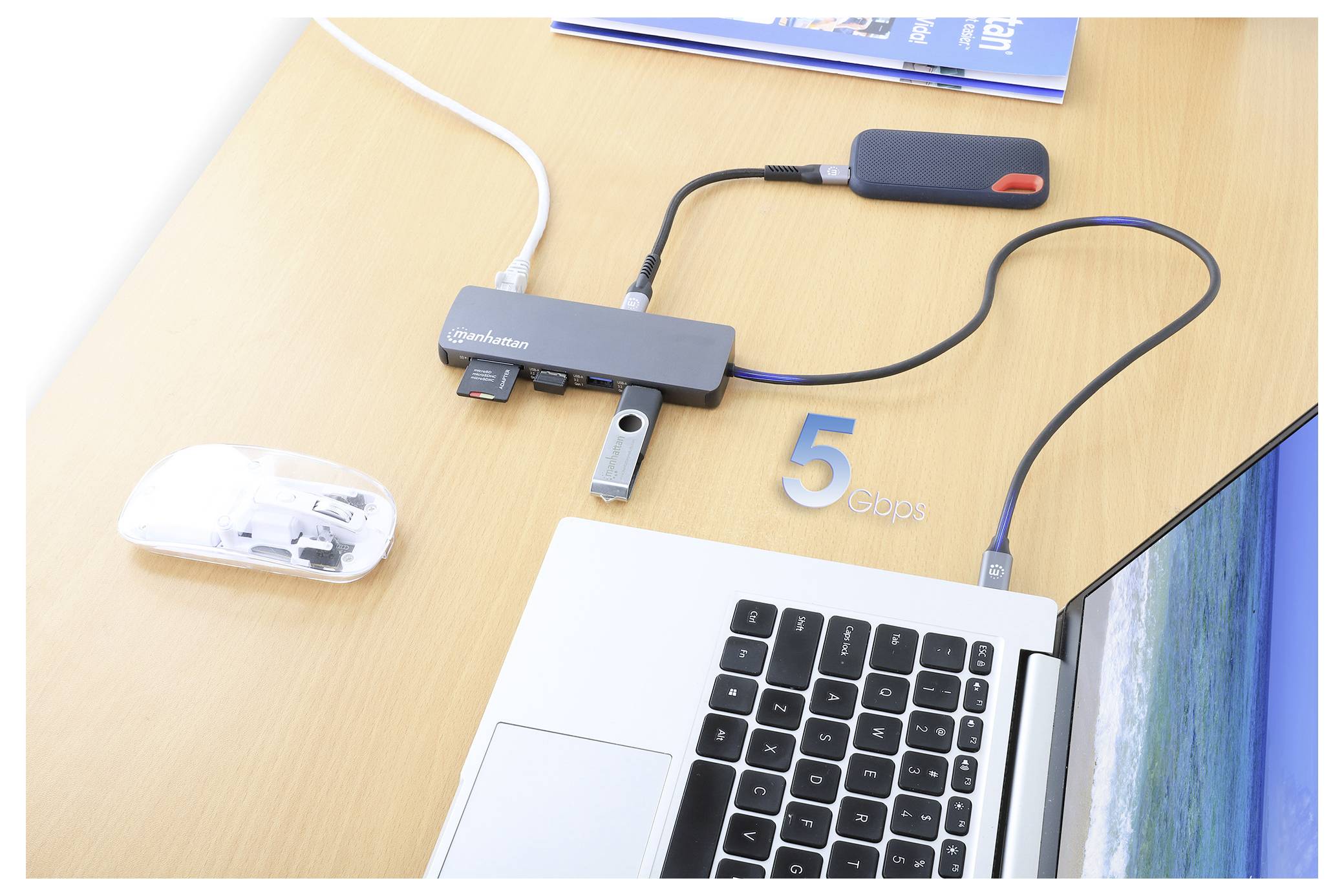 Ein Laptop ist über einen mit '5 Gbps' beschrifteten USB-Hub mit mehreren Geräten verbunden. In der Nähe befinden sich ein Buch, eine weiße Maus und eine externe Festplatte.