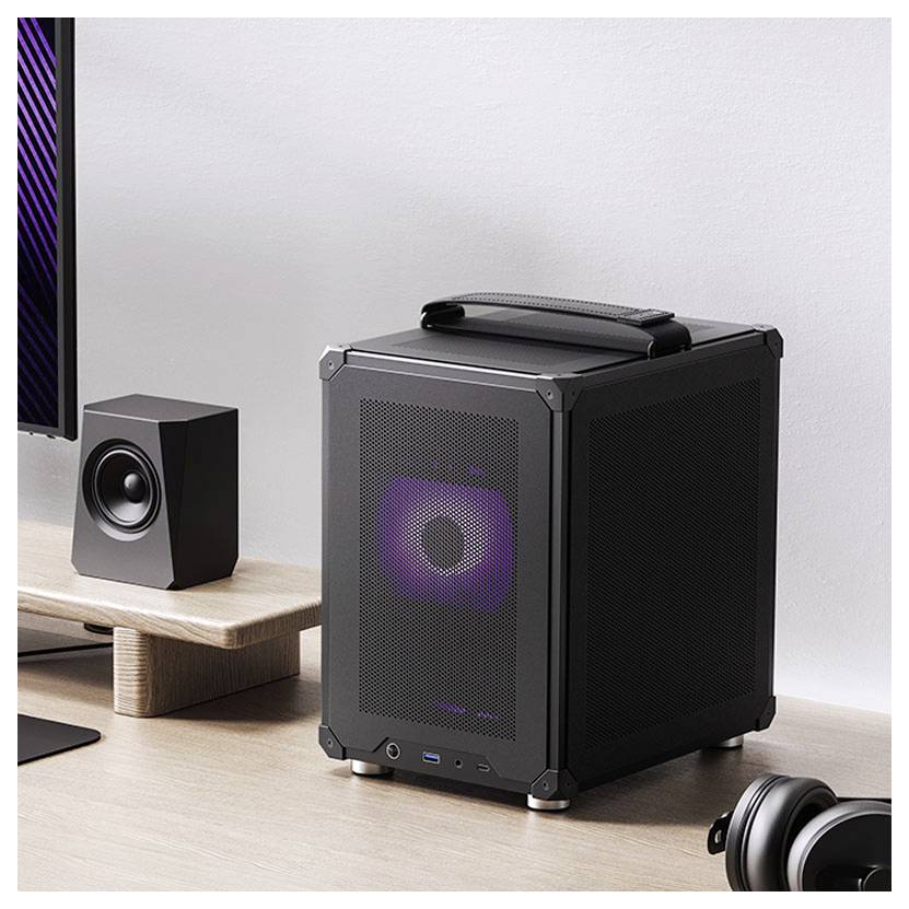 Ein kleiner, würfelförmiger Desktop-Computer mit Frontgitter und RGB-Beleuchtung steht auf einem Holzschreibtisch neben einem Lautsprecher.