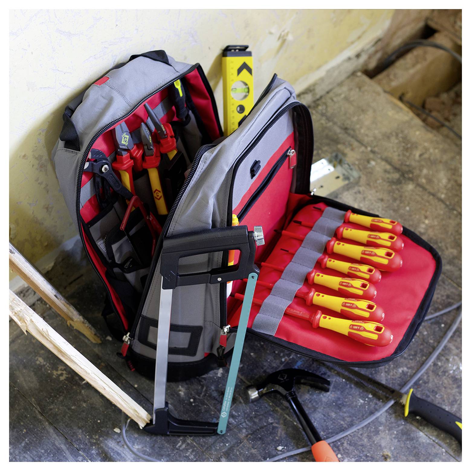 Eine offene Werkzeugtasche gefüllt mit verschiedenen Werkzeugen, darunter eine Handsäge, eine Wasserwaage, Schraubendreher und ein Hammer, die auf einem Holzboden stehen.