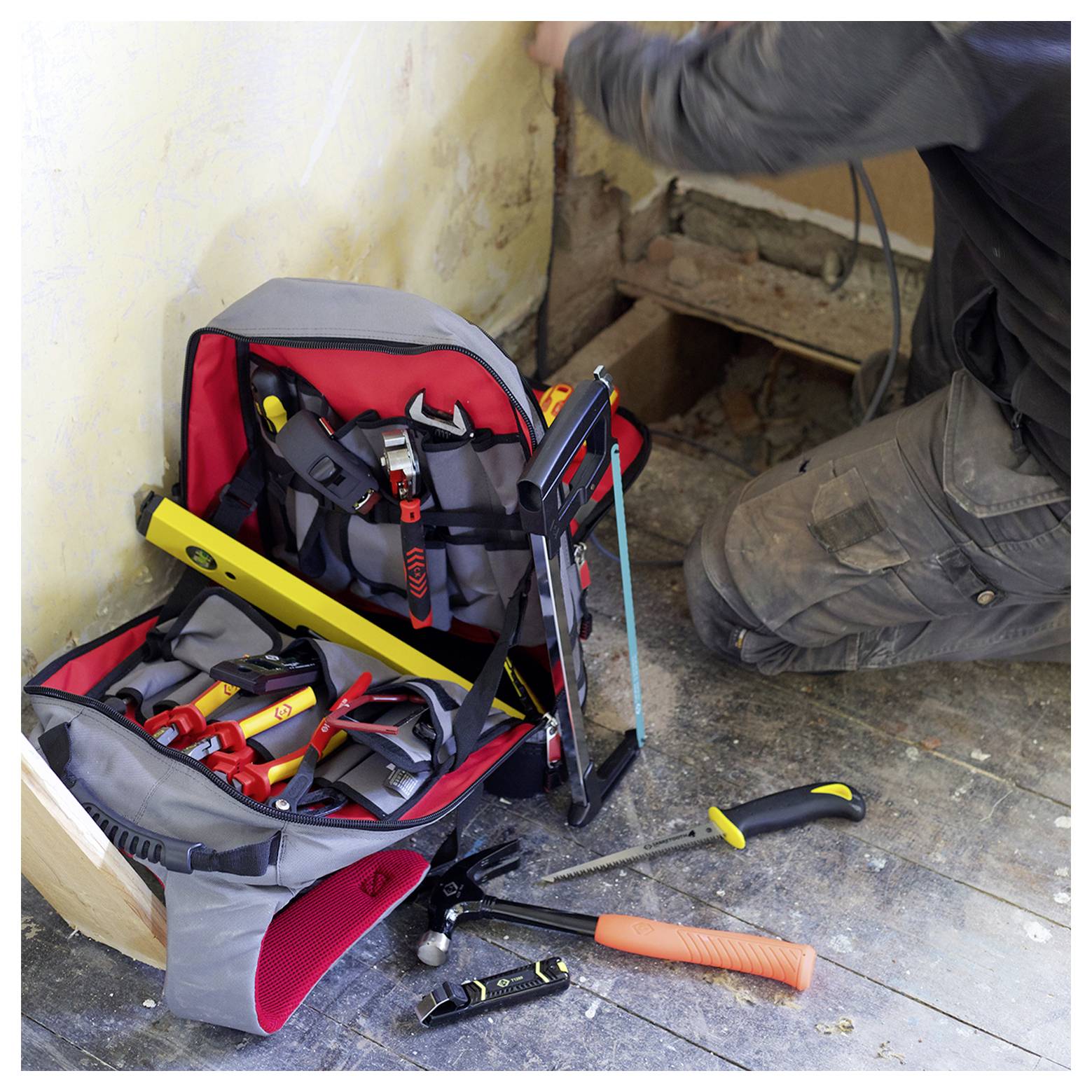 Eine Person kniet auf einem Holzboden in der Nähe einer Wand mit einer offenen Werkzeugtasche, die voller Werkzeuge wie Zangen, einer Wasserwaage und einer Säge ist, und diese werden gerade benutzt.