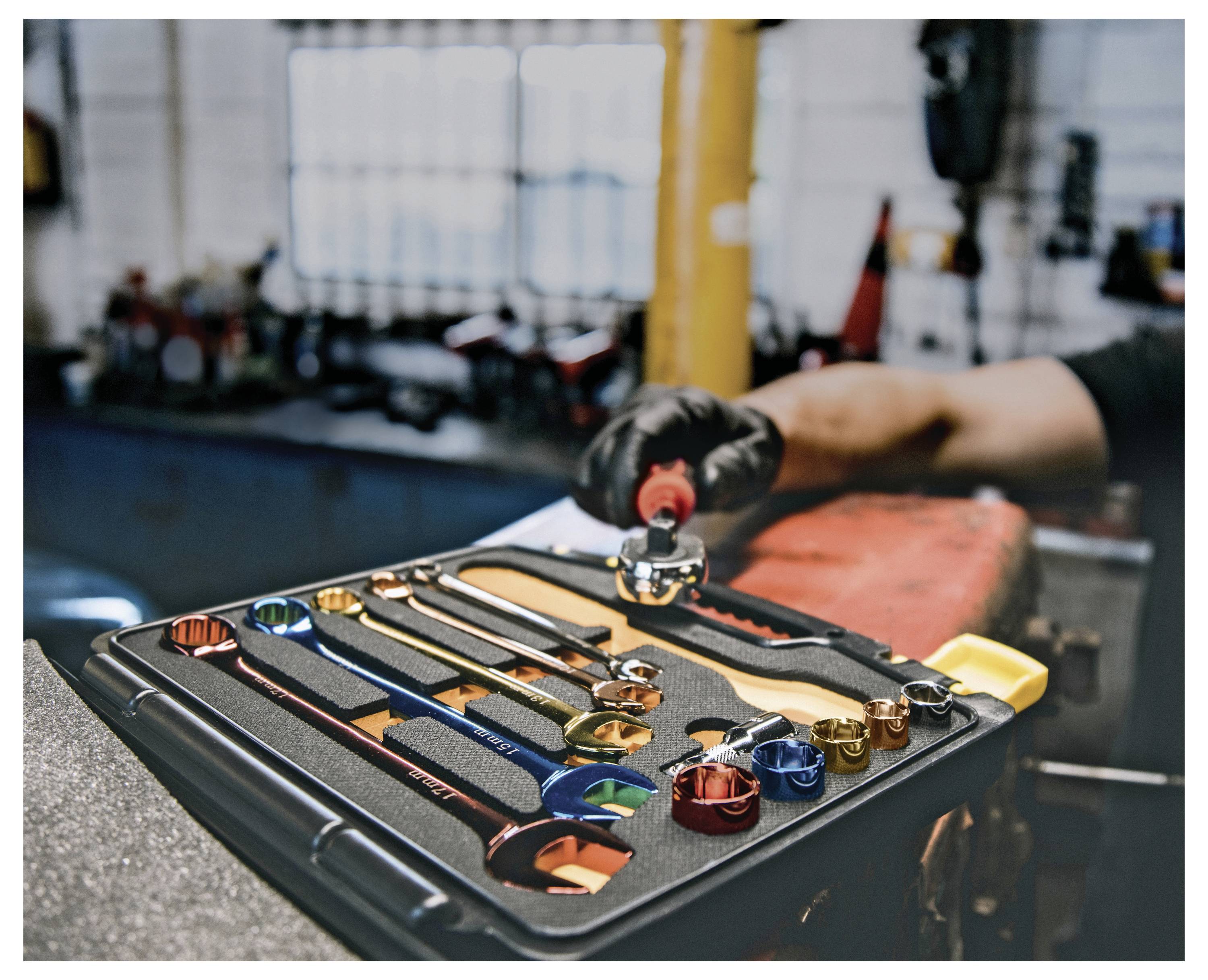 Eine Hand eines Mechanikers greift nach einem Werkzeug aus einer sauber organisierten Werkzeugkiste in einer Werkstatt. Verschiedene Schraubenschlüssel, Steckschlüssel und Ratschenschlüssel sind zu sehen.