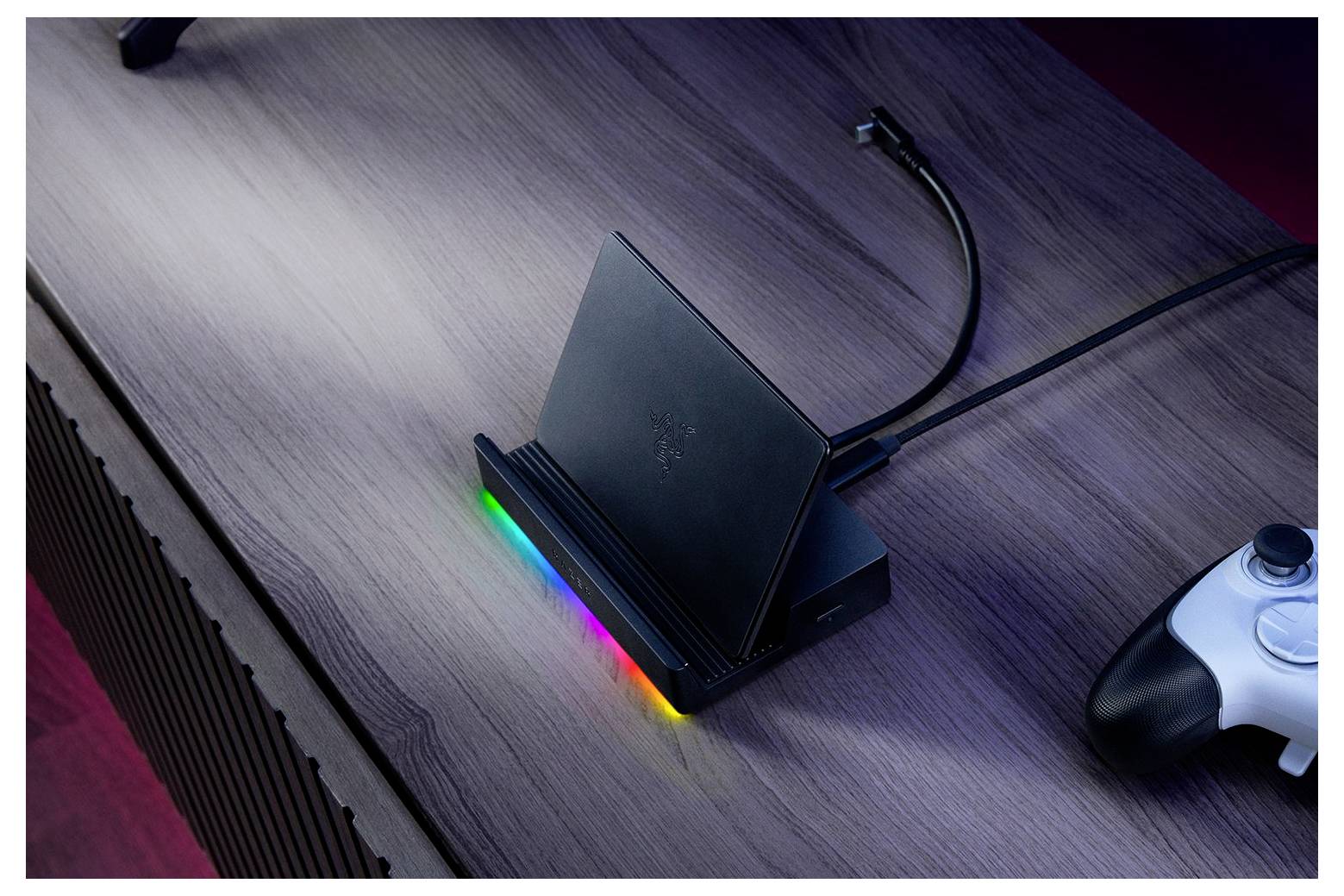 Eine Spielkonsole mit einer leuchtenden Rainbow-LED steht auf einem Holztisch. Ein Controller liegt in der Nähe und ist über ein Kabel verbunden.