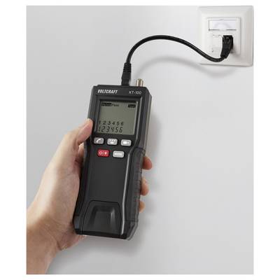 Eine Hand hält einen VOLTCRAFT KT-100 Kabeltester, der mit einer Wandsteckdose verbunden ist. Das Display zeigt Zahlen und das Gerät befindet sich im Testmodus.