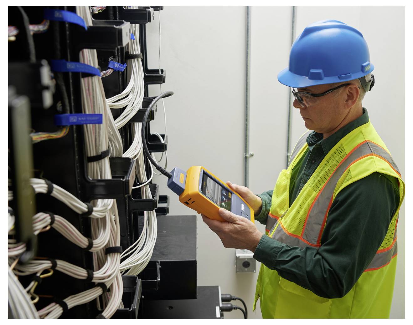 Ein Techniker in Schutzausrüstung untersucht die elektrische Verkabelung mit einem tragbaren Gerät in einem Kontrollraum und stellt die Systemfunktionalität sicher.