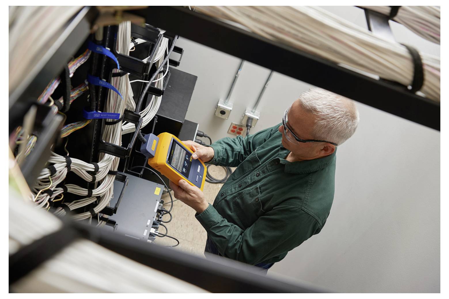 Eine Person, die in einer Serverraumumgebung ein Netzwerkpanel mit einem tragbaren elektronischen Messgerät überprüft, umgeben von Kabeln.