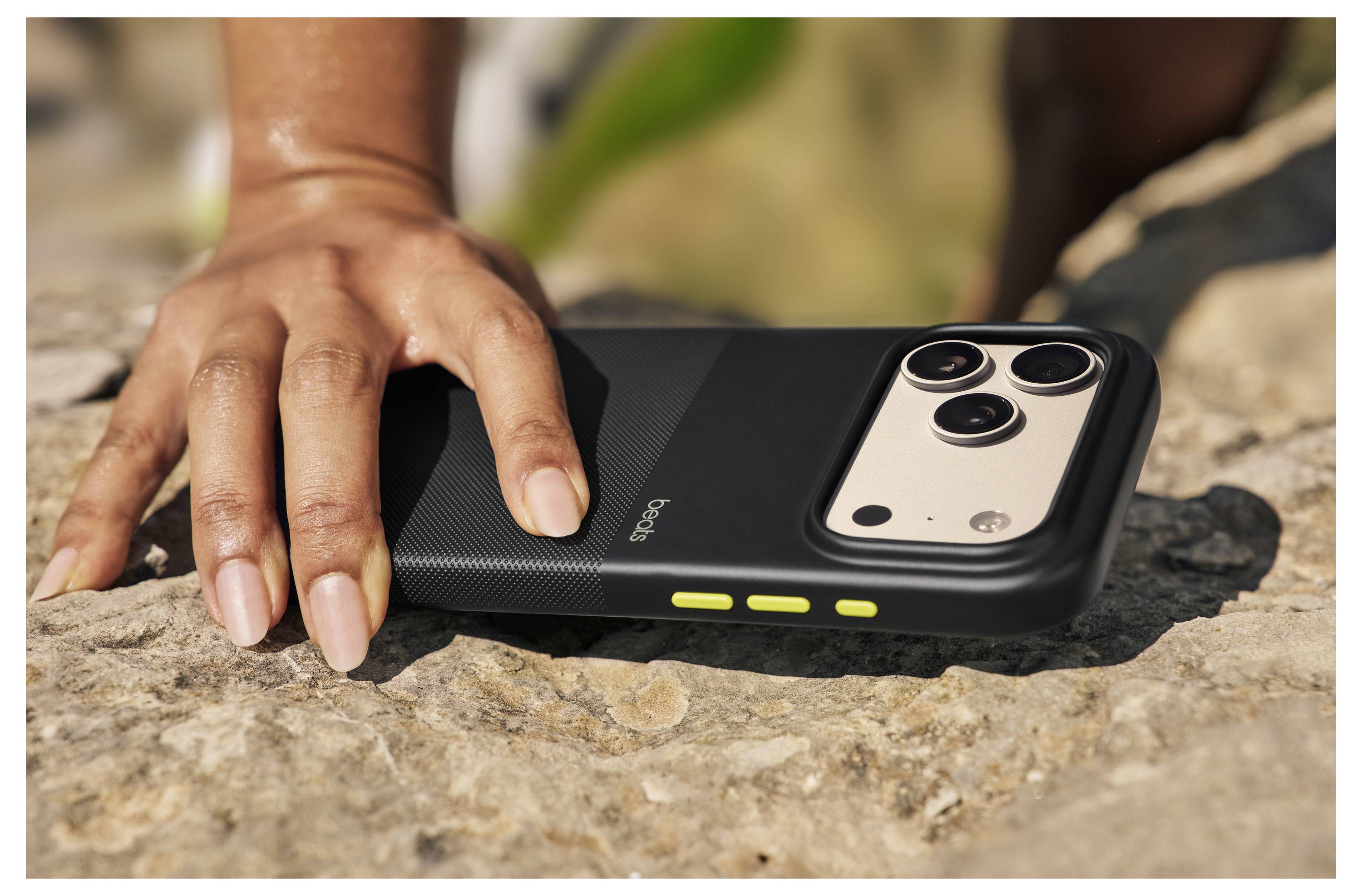 Eine Hand ruht auf einem Felsen, daneben liegt ein Smartphone in einem schwarz-goldenen Etui. Die Rückseite des Telefons verfügt über drei Kameraobjektive und gelbe Tasten.
