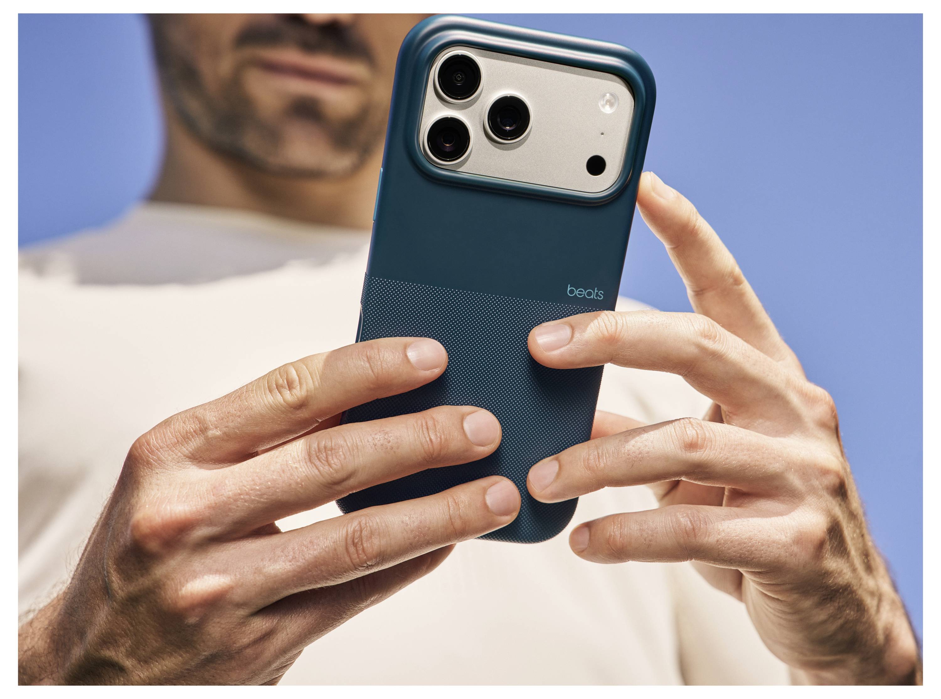 Eine Person in einem weißen Hemd hält ein Smartphone mit einem blauen Schutzgehäuse und einem hervorstehenden Kameramodul vor einem klaren blauen Himmelshintergrund.