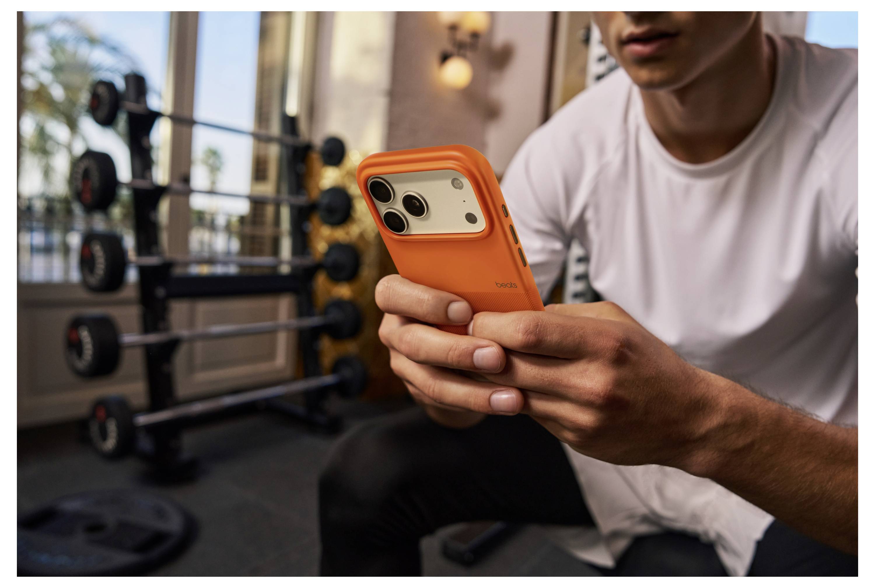 Eine Person in einer Gymumgebung hält ein Smartphone mit einer orangefarbenen Hülle, während sie in der Nähe von Fitnessgeräten wie einem Gewichteständer sitzt.