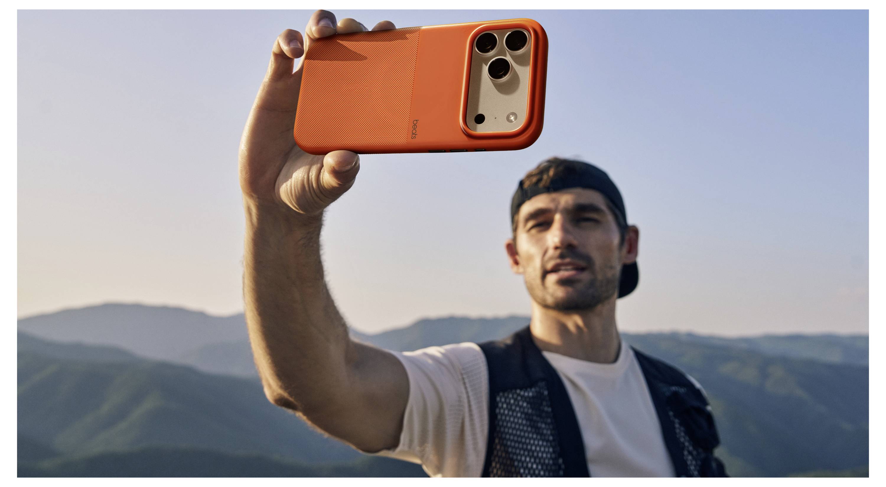Eine Person steht im Freien mit Bergen im Hintergrund und hält ein Smartphone in einem hellen orangefarbenen Etui für ein Selfie oder einen Videoanruf.