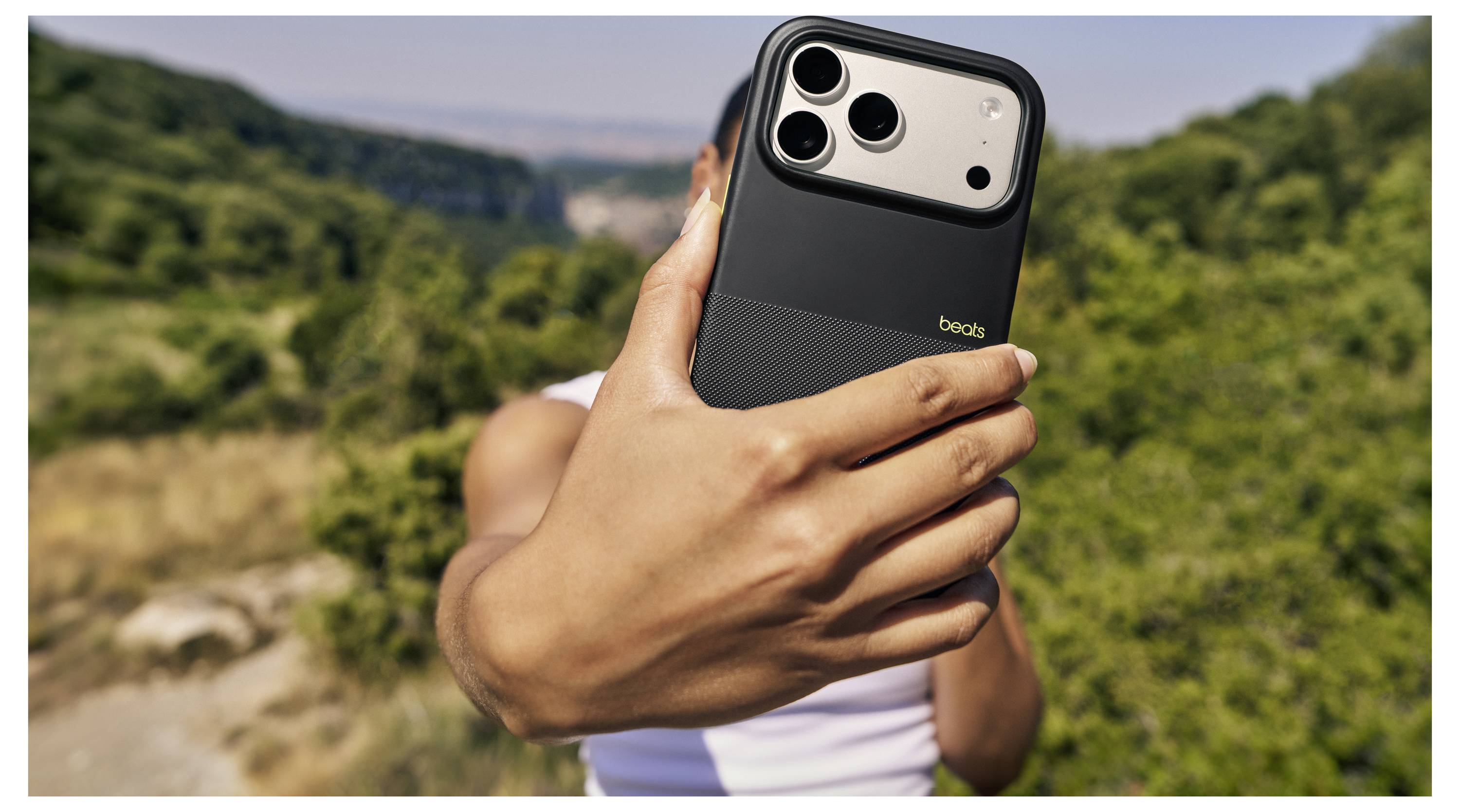 Eine Person hält ein Smartphone mit Dual-Objektiv-Kamera im Freien, wobei der Fokus auf dem Gerät vor einem unscharfen natürlichen Landschaftshintergrund liegt.