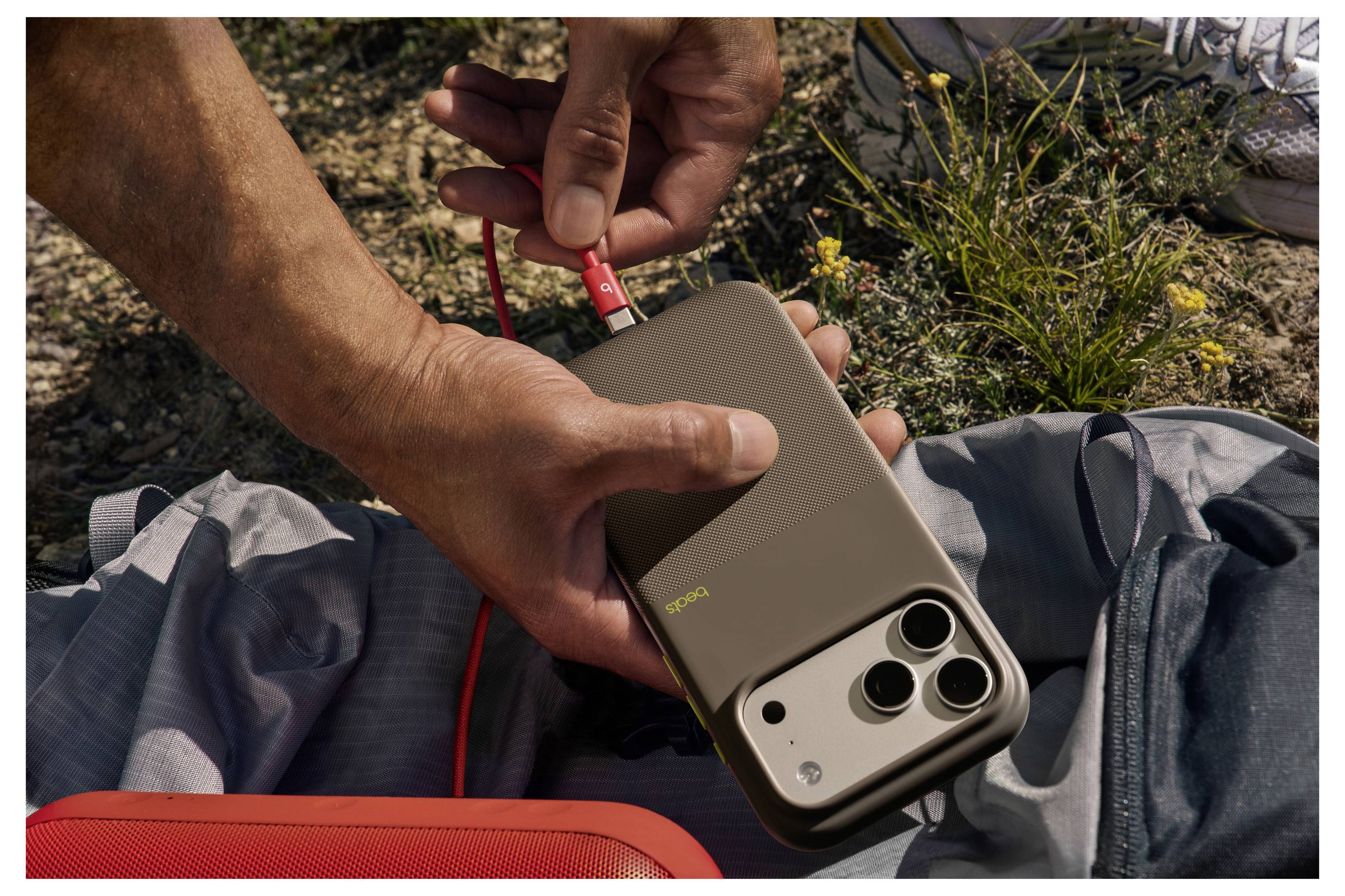 Eine Person lädt ein Smartphone mit einem orangefarbenen Kabel im Freien auf einem grauen Rucksack, wobei Gras und Erde in der Umgebung sichtbar sind.