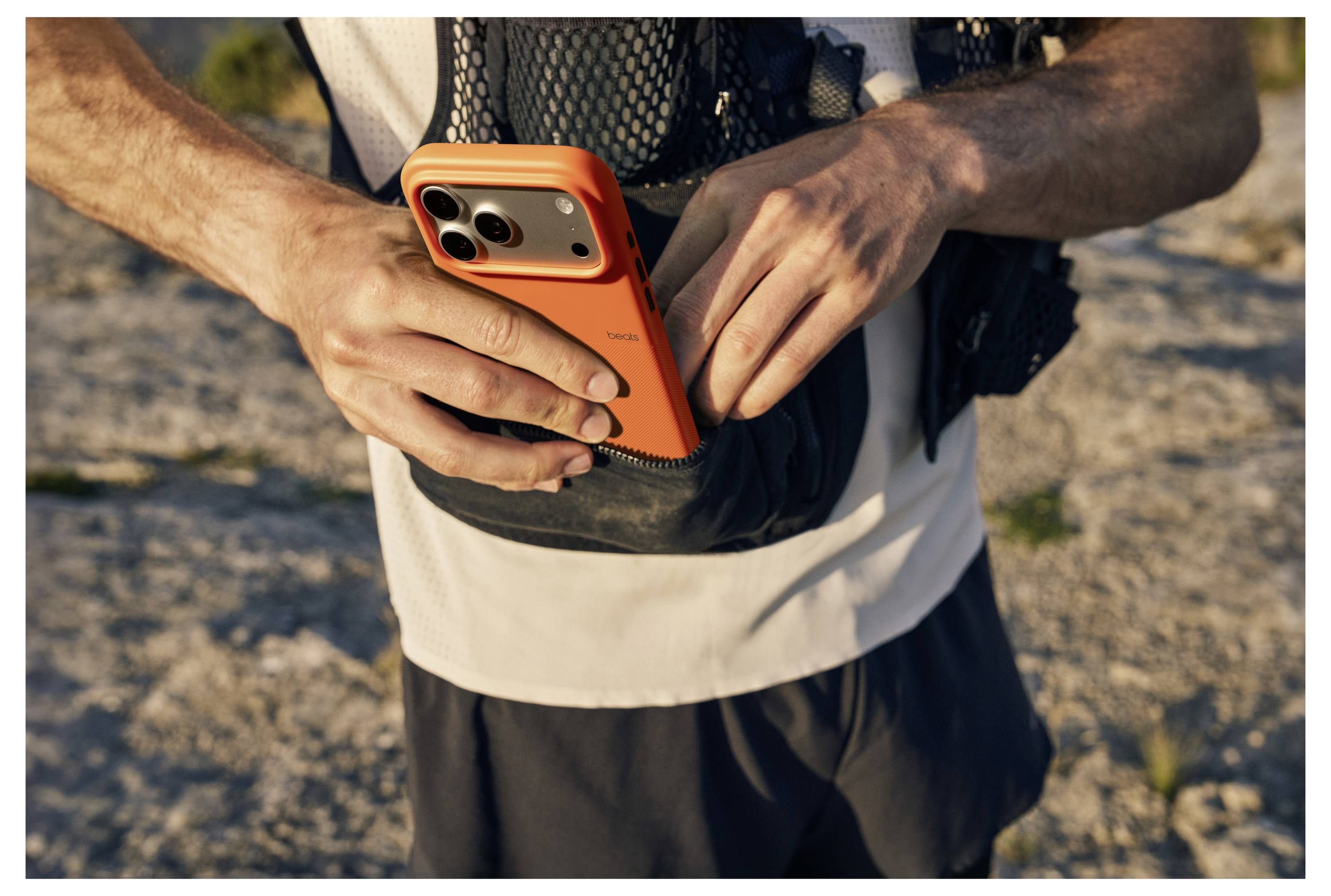 Eine Person legt ein orangefarbenes Smartphone in eine schwarze Tasche einer Netzweste und steht dabei im Freien auf felsigem Gelände, was auf eine Outdoor-Aktivität hindeutet.