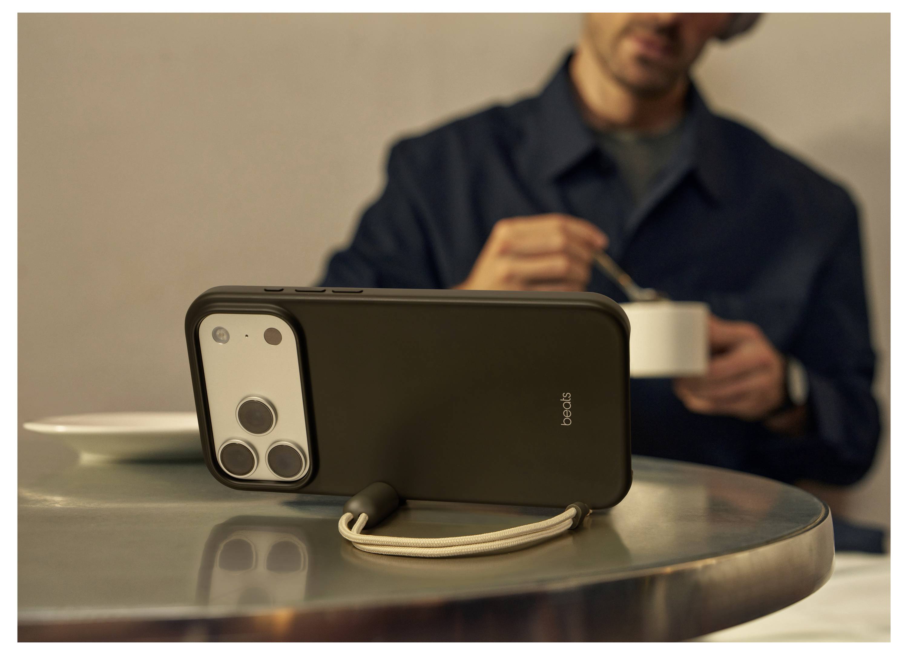 Ein Smartphone mit einer Schutzhülle liegt im Vordergrund auf einem Tisch, während eine Person im Hintergrund konzentriert aus einer Schüssel isst.