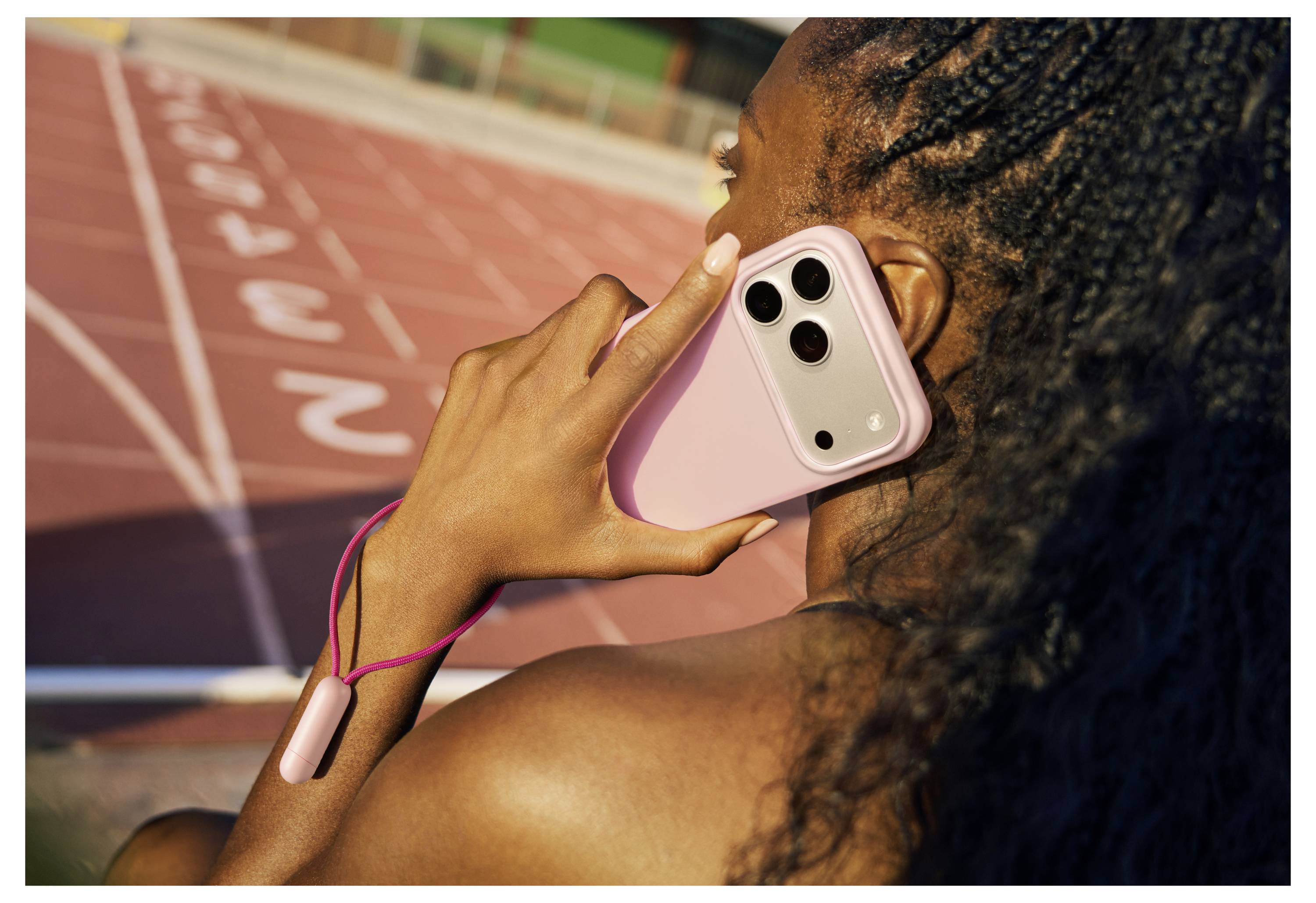 Eine Person hält ein rosa Smartphone an ihr Ohr, während sie sich auf einer Laufbahn mit sichtbaren Bahnen befindet.