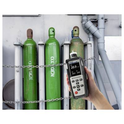 Eine Hand hält ein Gasprüfgerät mit einer Anzeige von „20,9" in der Nähe von geketteten grünen Sauerstofftanks, was auf einen sicheren Sauerstoffgehalt hinweist.