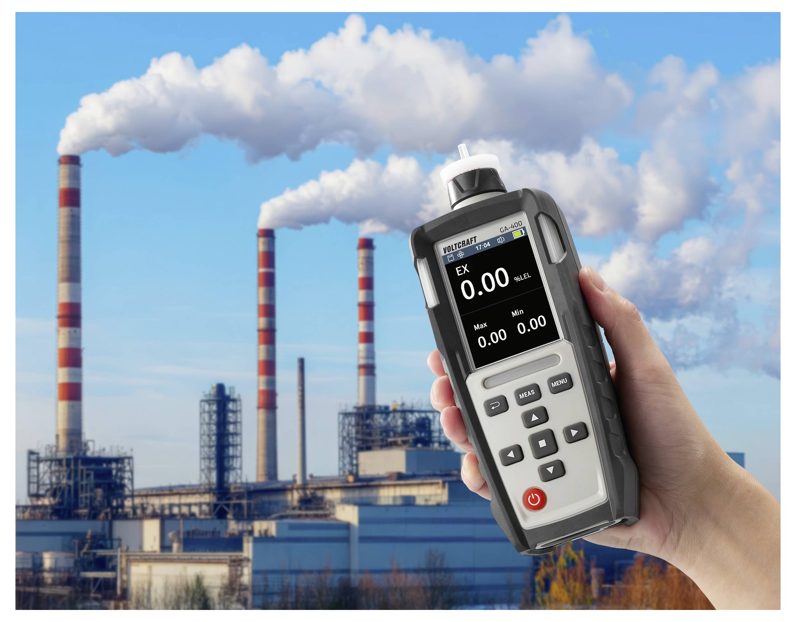 Eine Hand hält einen tragbaren Gasdetektor mit einer Anzeige von „0,00"; Industrieschornsteine im Hintergrund stoßen Rauch unter blauem Himmel aus.