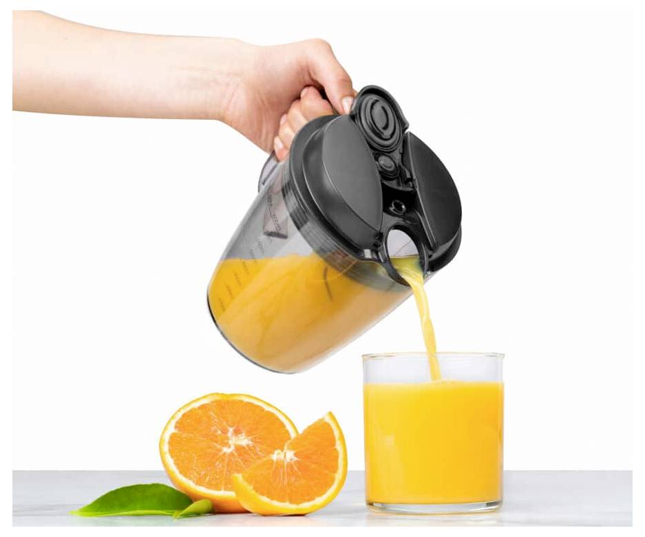 Eine Hand gießt Orangensaft aus einem Krug in ein Glas auf einer weißen Oberfläche. Frische Orangenscheiben und Blätter liegen neben dem Glas.