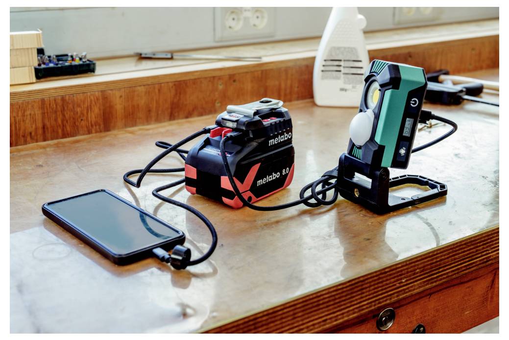 Ein Smartphone, ein Ladegerät mit der Aufschrift „metabo 18V" und eine tragbare Lampe liegen auf einer Holzwerkbank in der Nähe von Steckdosen.