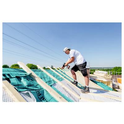 Ein Bauarbeiter in einem weißen Hemd und kurzen Hosen installiert Dachbalken auf einem Dach unter einem klaren blauen Himmel.