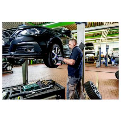 Ein Mechaniker in einer Werkstatt inspiziert das Reifen eines Autos auf einer Hebebühne. Werkzeuge und Ausrüstung sind im Hintergrund der Werkstatt sichtbar.