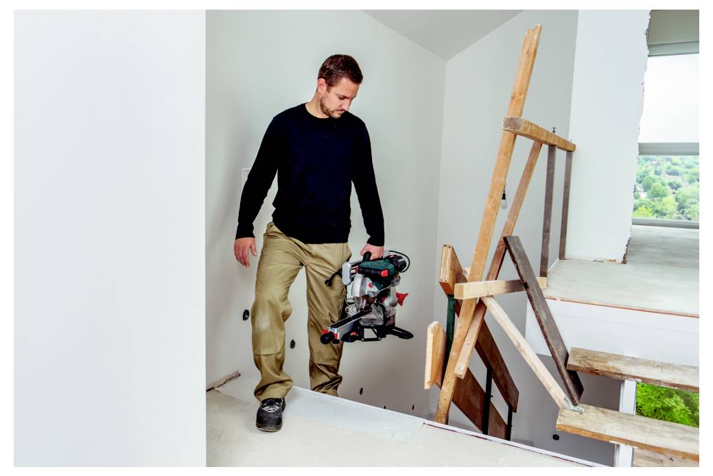 Ein Mann in einem dunklen langärmligen Hemd und beigen Hosen steht auf einer unfertigen Treppe und hält ein Elektrowerkzeug in einem Gebäude im Bau.