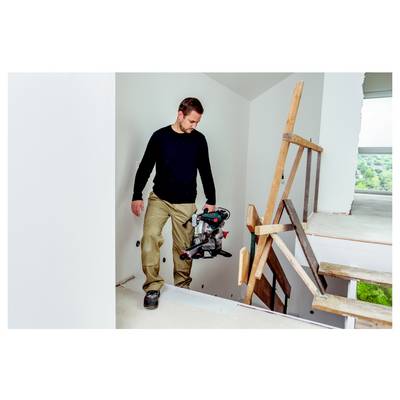 Ein Mann in einem dunklen langärmligen Hemd und beigen Hosen steht auf einer unfertigen Treppe und hält ein Elektrowerkzeug in einem Gebäude im Bau.