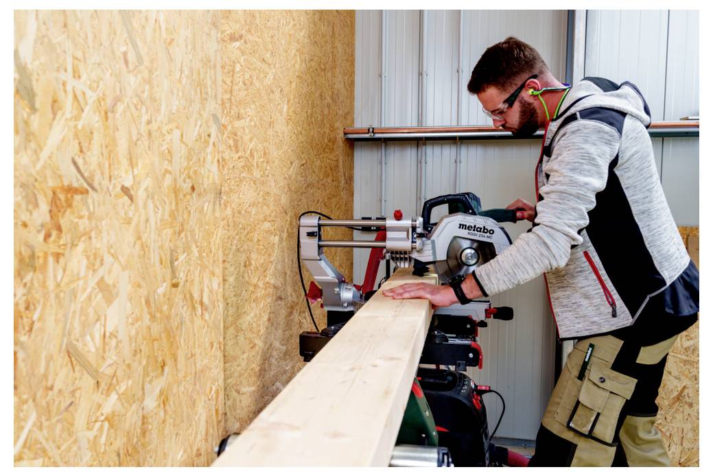 Eine Person bedient eine Gehrungssäge, die ein Holzbrett in einer Werkstatt zersägt. Sie trägt Sicherheitsbrille und Gehörschutz und konzentriert sich auf Präzision.