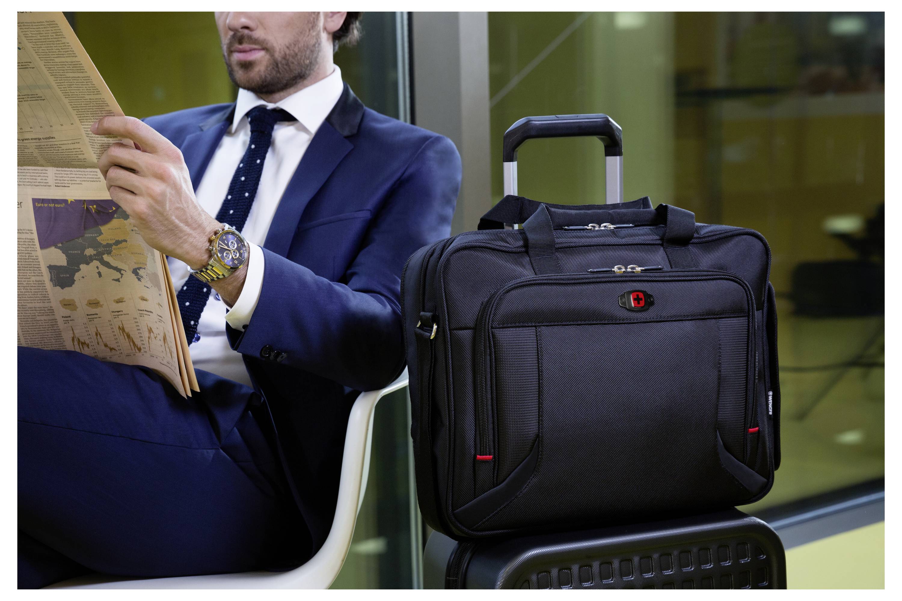 Mann im Anzug, der eine Zeitung liest, sitzt nahe einem Fenster, mit einem schwarzen Aktenkoffer auf einem Rollkoffer neben sich, was Reise oder Geschäftlichkeit andeutet.