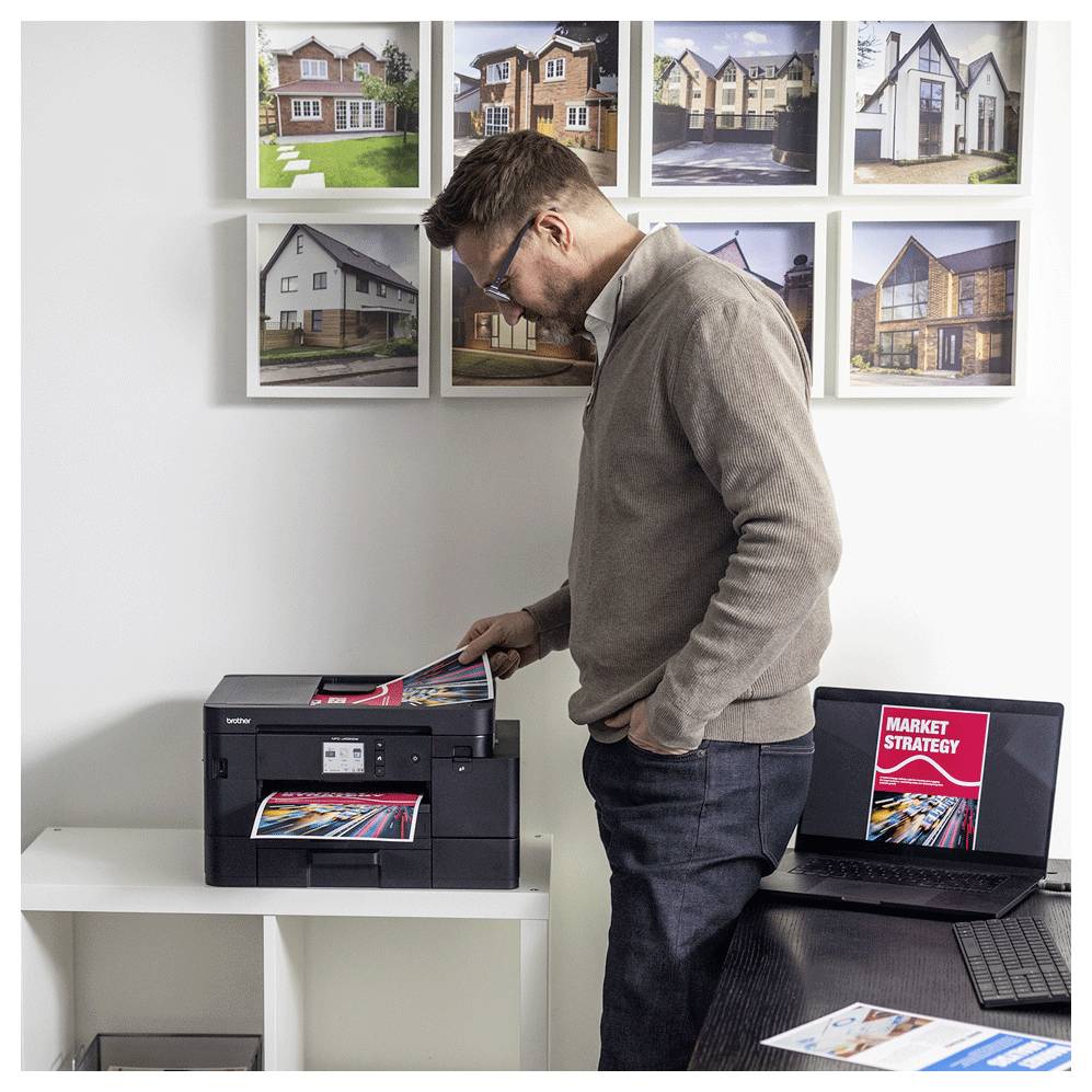 Ein Mann bedient einen Drucker in einem Büro, umgeben von gerahmten Fotos von Häusern. Ein Laptop zeigt ein Dokument mit dem Titel "Marktstrategien".