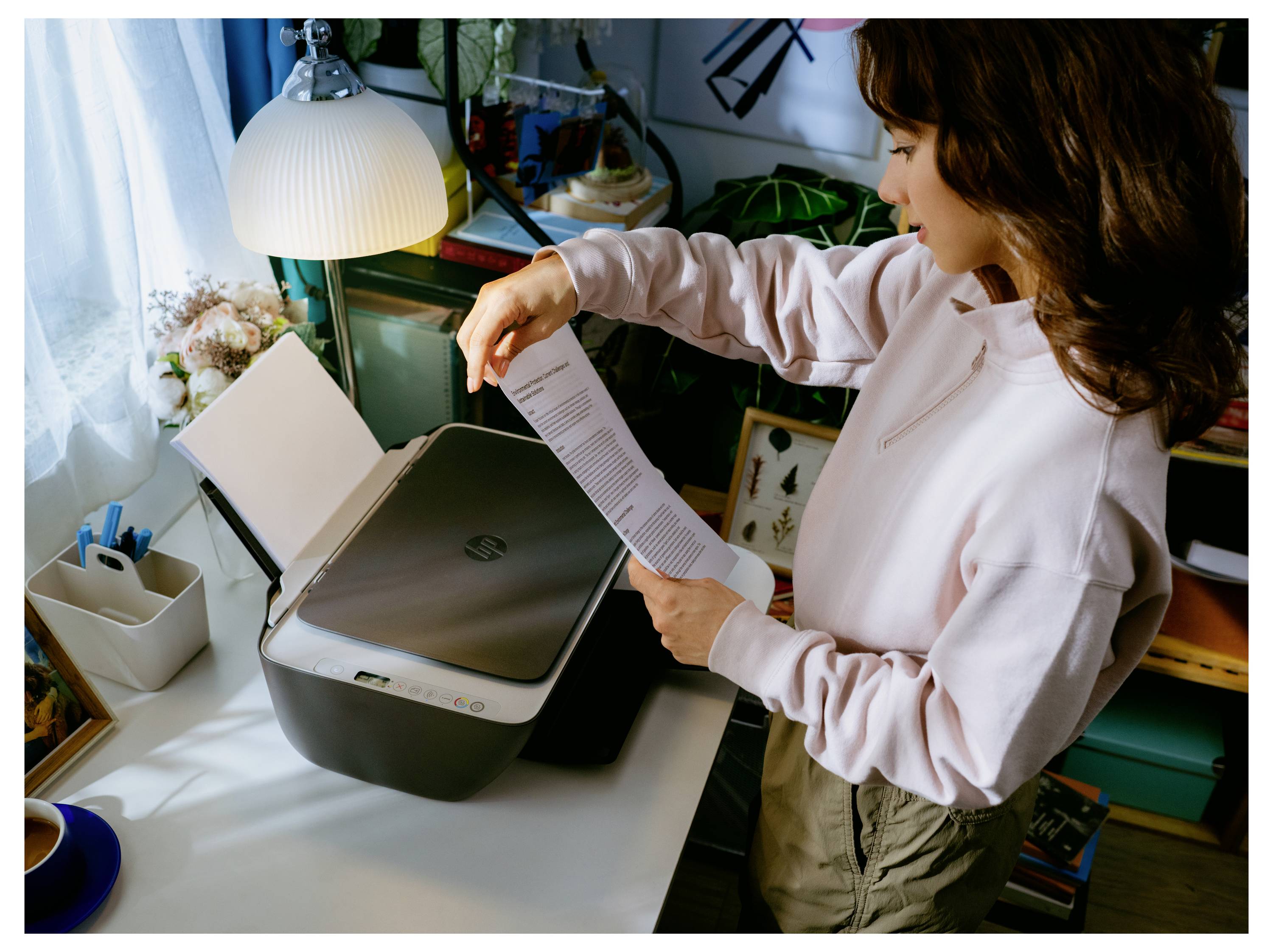 Frau überprüft ein Dokument, während sie einen Flachbettscanner auf einem Schreibtisch zu Hause benutzt, mit einer Lampe und Büromaterialien in der Nähe.