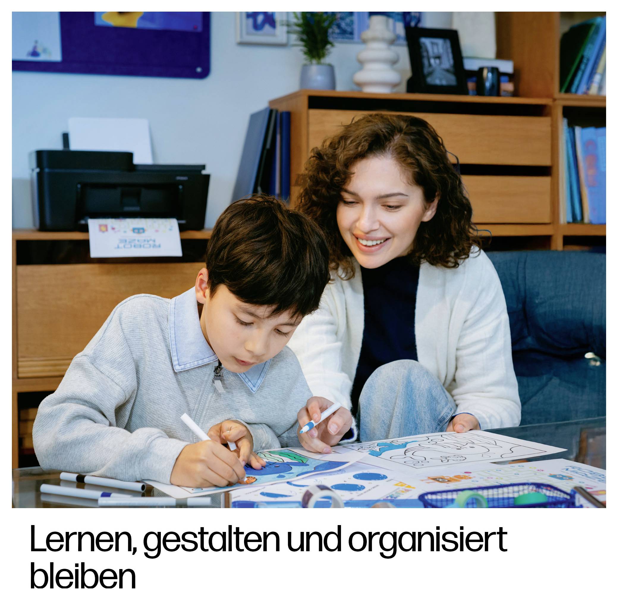 # Lernen, gestalten und organisiert bleiben<br><br>Eine Frau hilft einem Jungen, auf Papieren zu malen und zu zeichnen; der deutsche Text darunter lautet „Lernen, gestalten und organisiert bleiben"