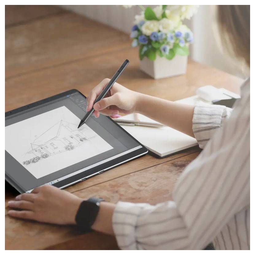 Eine Person verwendet einen Stylus, um ein Haus auf einem Tablet zu zeichnen. Ein Notizbuch und Topfblumen stehen im Hintergrund auf dem Holzschreibtisch.