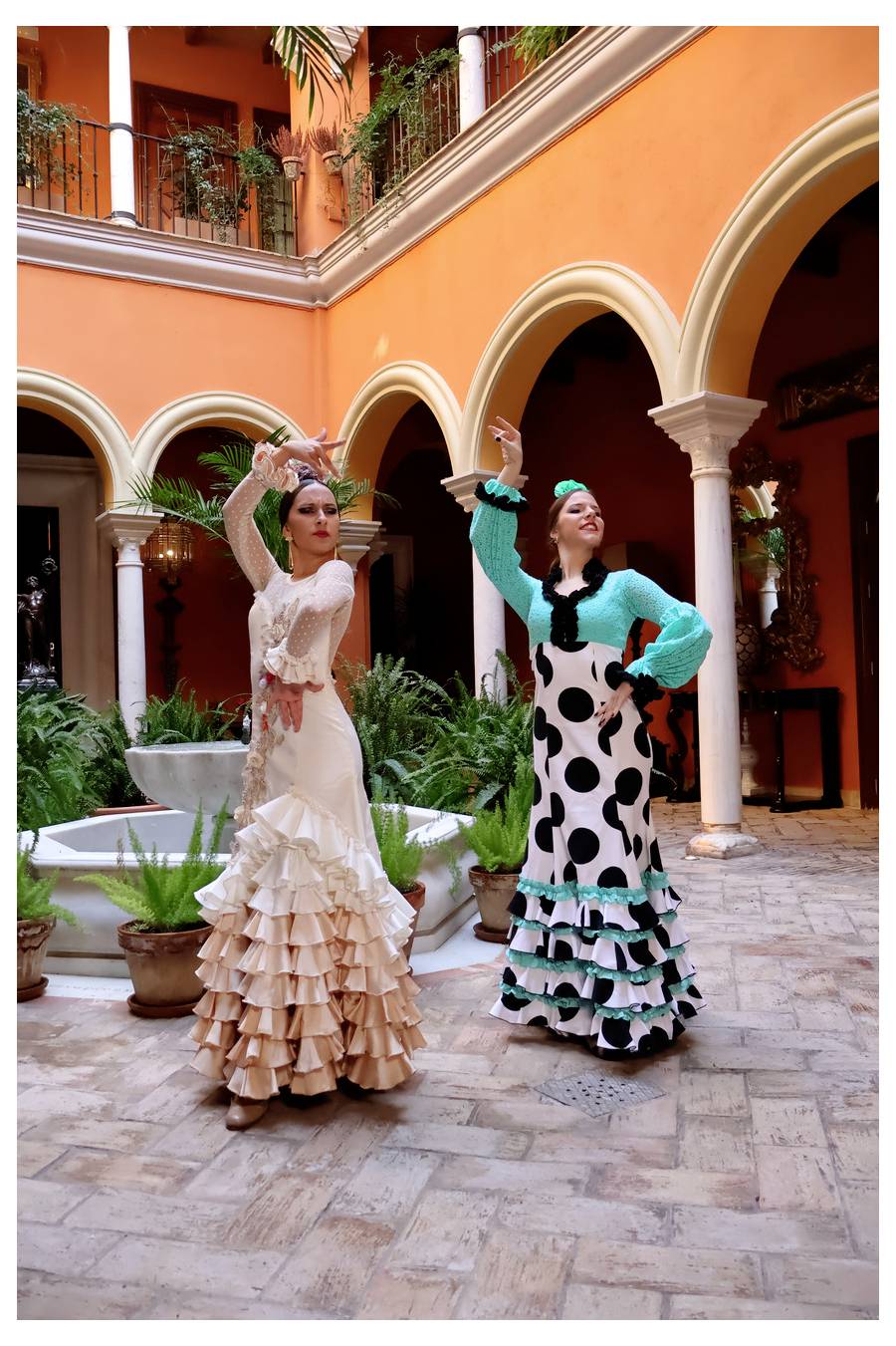 Zwei Tänzerinnen in traditionellen spanischen Flamenco-Kleidern posieren mit erhobenen Armen in einem lebhaften Innenhof, umgeben von Bögen und Topfpflanzen.