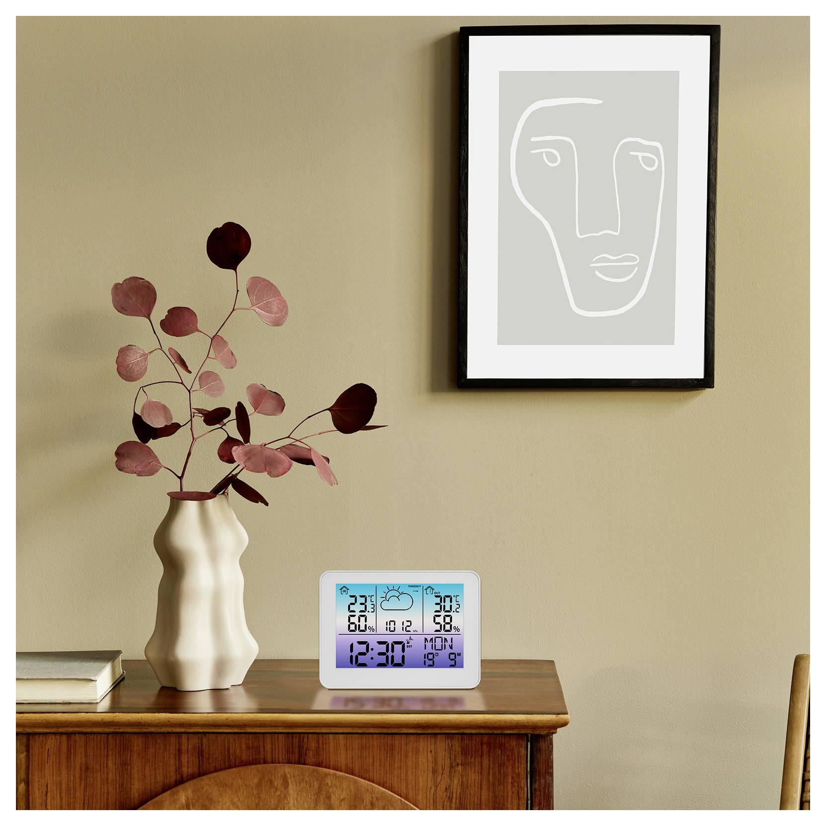 Eine digitale Uhr auf einem Holztisch zeigt '12:30' mit zusätzlichen Informationen wie Datum, Temperatur und Luftfeuchtigkeit. In der Nähe steht eine Vase mit Zweigen, und an der Wand hängt ein gerahmtes abstraktes Gesichtskunstwerk.